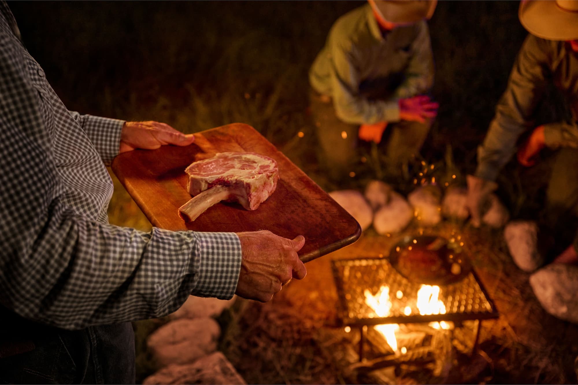 A person holds a raw tomahawk steak on a wooden cutting board near an outdoor campfire at night, with two other people visible in the background.