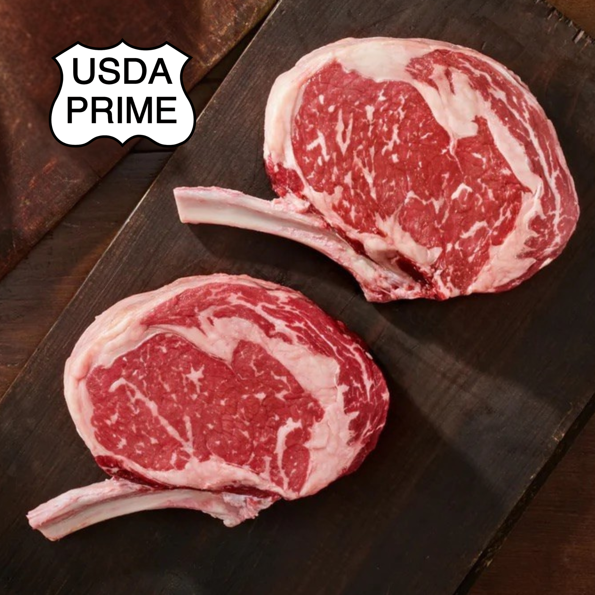 Two USDA Prime ribeye steaks with bone-in bones, displayed on a dark wooden cutting board with a USDA Prime logo badge in the top left corner.